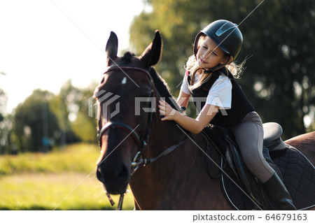 Girl teenager jockey sits on a brown horse, hugs and strokes her. Girl teenager jockey sits on a brown horse, hugs and strokes her. 64679163
