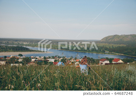 a little girl in a loose dress with loose hair walks in a meadow. Village houses, forest and river as background. The concept of summer, warmth, freedom, village life, sunburn, chldhood 64687248