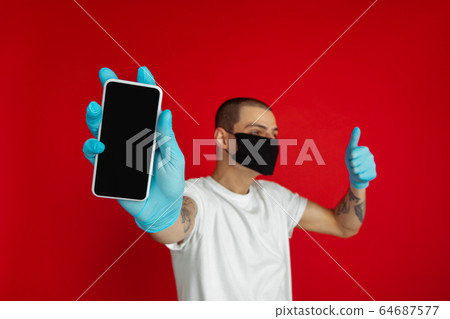 Caucasian young man in protective face mask and medical gloves on red studio background - showing blank phone's screen Caucasian young man in protective face mask and medical gloves on red studio background - showing blank phone's screen 64687577
