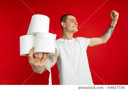 Caucasian young man's portrait on red studio background - holding toilet papers, essential goods during quarantine and self-insulation Caucasian young man's portrait on red studio background - holding toilet papers, essential goods during quarantine and self-insulation 64687596