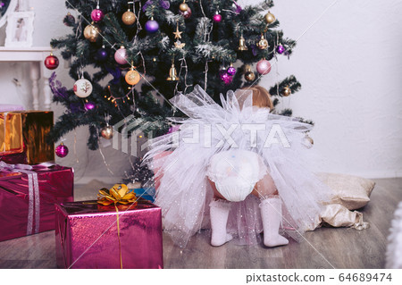 Little girl in a beautiful dress and a diaper near the New Year tree Little girl in a beautiful dress and a diaper near the New Year tree 64689474