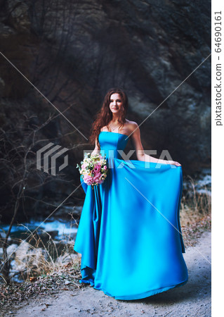 The brunette at the foot of the mountain stands in a blue dress with a bouquet of flowers The brunette at the foot of the mountain stands in a blue dress with a bouquet of flowers 64690161