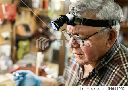 Senior craftsman portrait with headlamp led torch working in workhop with tools and equipment on background. Old mature gray handyman tutor with eyeglasses. Experinced person making diy hobby craft 64691062