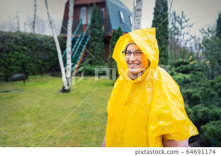 Portrait of beautiful young adult caucasian woman under bright yellow raincoat and wellies walking at garden during cold spring rainy day at home backyard. Carefree hipster concept. Bad weather Portrait of beautiful young adult caucasian woman under bright yellow raincoat and wellies walking at garden during cold spring rainy day at home backyard. Carefree hipster concept. Bad weather 64691174