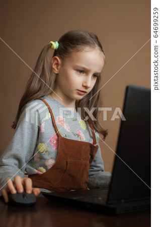 Serious little girl is concentrated in front of her black laptop while focused on doing homework during the quarantine because of the pandemia of Coronavirus Covid-19. Remote education Serious little girl is concentrated in front of her black laptop while focused on doing homework during the quarantine because of the pandemia of Coronavirus Covid-19. Remote education 64699259