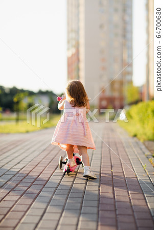 Little girl wearing dress riding scooter outdoors Little girl wearing dress riding scooter outdoors 64700680