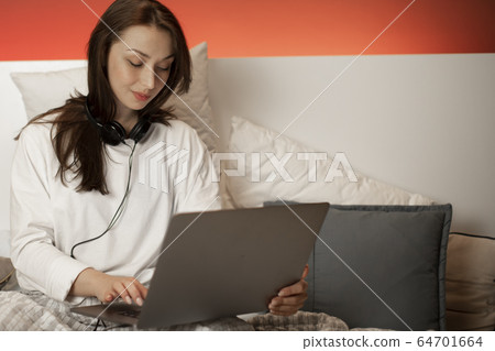 Young beautiful girl in headphones sitting in bed, works on laptop from home Young beautiful girl in headphones sitting in bed, works on laptop from home 64701664