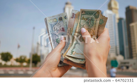 The girl holds in her hands the money of the United Arab Emirates on the background of the city center of Dubai. 64702331