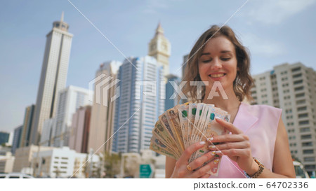 The girl holds in her hands the money of the United Arab Emirates on the background of the city center of Dubai. 64702336