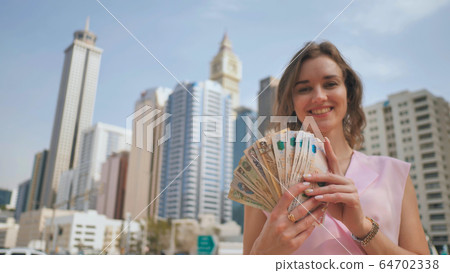 The girl holds in her hands the money of the United Arab Emirates on the background of the city center of Dubai. 64702338