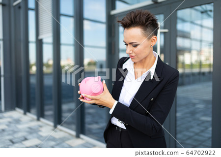 Caucasian woman in office clothes holds a pink pig moneybox and waits a coleague near the office 64702922