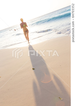 Happy woman enjoying in summer, running joyfully on tropical beach in sunset. Beautiful caucasian model wearing bikini on vacations on sandy beach. Footprints in sand. Copy space 64703352