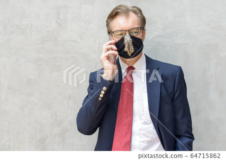 Mature businessman wearing mask and talking on the phone against concrete wall Mature businessman wearing mask and talking on the phone against concrete wall 64715862