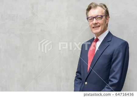 Handsome mature businessman in suit smiling and wearing eyeglasses against concrete wall 64716436
