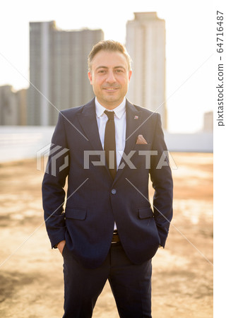 Happy mature handsome businessman in suit against view of the city 64716487