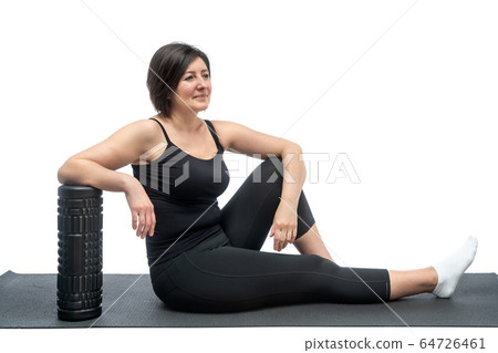 A middle-aged woman, on a gymnastic mat with myofascial roller, is resting after exercise on a white background. A middle-aged woman, on a gymnastic mat with myofascial roller, is resting after exercise on a white background. 64726461