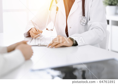 Unknown woman-doctor and female patient sitting and talking at medical examination in sunny clinic, close-up. Therapist filling up medication history record. Medicine concept Unknown woman-doctor and female patient sitting and talking at medical examination in sunny clinic, close-up. Therapist filling up medication history record. Medicine concept 64761891