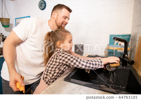 Happy young father standing by his little daughter and helping her to wash cup 64763185