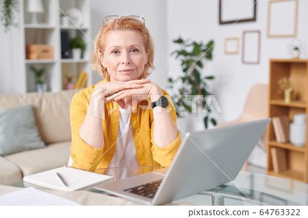 Pretty blond businesswoman in yellow shirt looking at you while working remotely Pretty blond businesswoman in yellow shirt looking at you while working remotely 64763322