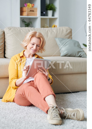 Mature relaxed businesswoman with mobile gadget sitting on the floor by couch 64763349