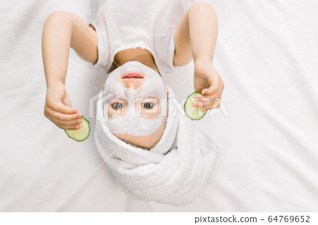 Funny little girl with a mask for skin face and towel on her head, holding two slices of fresh cucumber and looking at camera, while lying on white background after bath Funny little girl with a mask for skin face and towel on her head, holding two slices of fresh cucumber and looking at camera, while lying on white background after bath 64769652