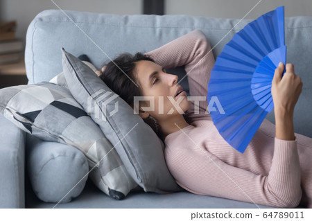 Unhealthy millennial girl cooling herself with paper fun indoors. 64789011