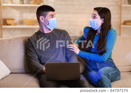 Scared couple in face masks using laptop sitting in sofa 64797374