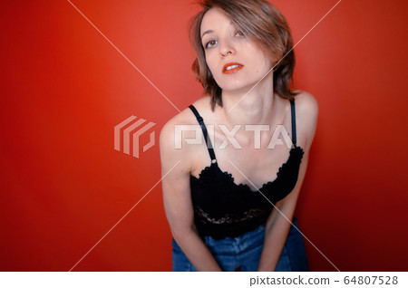 Young woman with short hair dressed in black lace bustier and blue jeans is posing in the studio on bright red background 64807528