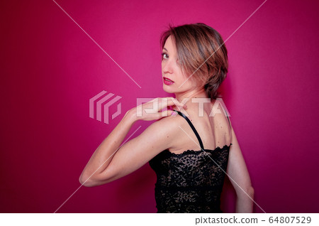 Young woman with short hair dressed in black lace bustier and blue jeans is posing in the studio on bright pink background 64807529