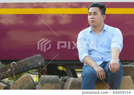 Young overweight Asian tourist man thinking by the train at railway train station Young overweight Asian tourist man thinking by the train at railway train station 64809554