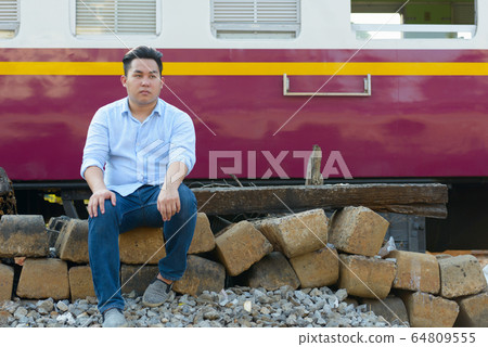 Young overweight Asian tourist man sitting by the train at railway train station Young overweight Asian tourist man sitting by the train at railway train station 64809555