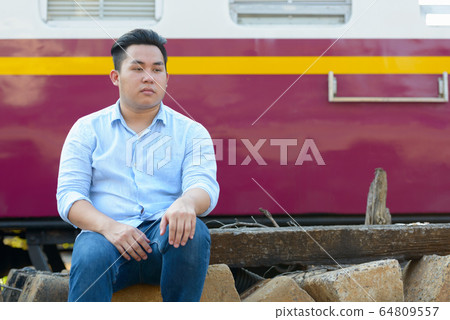 Young overweight Asian tourist man sitting by the train at railway train station Young overweight Asian tourist man sitting by the train at railway train station 64809557