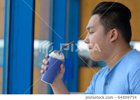 Young overweight Asian man drinking coffee at the coffee shop Young overweight Asian man drinking coffee at the coffee shop 64809754