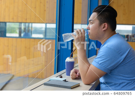 Young overweight Asian man drinking water at the coffee shop Young overweight Asian man drinking water at the coffee shop 64809765
