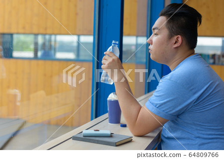 Young overweight Asian man drinking water at the coffee shop Young overweight Asian man drinking water at the coffee shop 64809766
