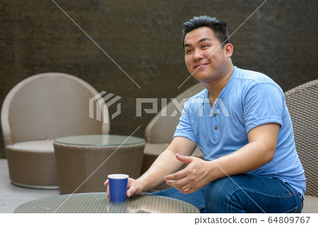 Happy young overweight Asian man drinking coffee at the coffee shop Happy young overweight Asian man drinking coffee at the coffee shop 64809767