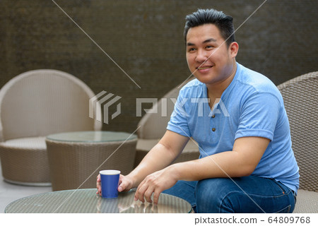 Happy young overweight Asian man drinking coffee at the coffee shop Happy young overweight Asian man drinking coffee at the coffee shop 64809768