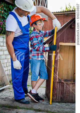 Carpenter watching his young trainee working with building level near wooden dog house at backyard of their house. Carpenter watching his young trainee working with building level near wooden dog house at backyard of their house. 64810807