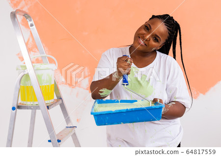 Young happy african american woman painting interior wall with paint roller in new house. A woman with roller applying paint on a wall. 64817559