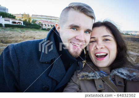 Portrait of young beautiful couple taking selfie photo in a smartphone with sea and dark cloudy sky on the background. Cold season and travel concept. Portrait of young beautiful couple taking selfie photo in a smartphone with sea and dark cloudy sky on the background. Cold season and travel concept. 64832302