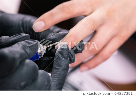 Hardware manicure in a beauty salon. Female manicurist is applying electric nail file drill to manicure on female fingers. Mechanical manicure close-up. Concept body care. 64837391