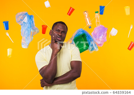 Problem of trash, plastic recycling, pollution and environmental concept - Serious african american man looking on trash on yellow background. He is thinking about ecology 64839164