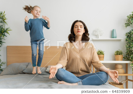 Mother meditating and her kid jumping on bed 64839909