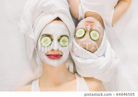 Mother and little daughter having spa procedures together. They are in white bath towels on head and with slices of cucumber on their eyes. Woman has white facial mask on her skin Mother and little daughter having spa procedures together. They are in white bath towels on head and with slices of cucumber on their eyes. Woman has white facial mask on her skin 64846505