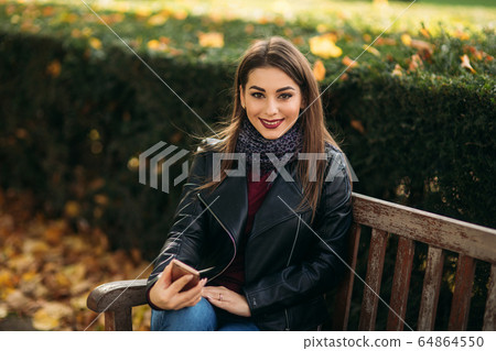 Stylish beautiful lady in a black leather jacket with a black bag and burgundy blouse. Attractive young woman use phone Stylish beautiful lady in a black leather jacket with a black bag and burgundy blouse. Attractive young woman use phone 64864550