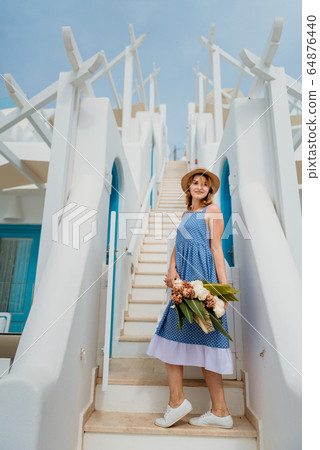 Beautiful girl walking with bouquet of flowers along the street of Santorini island, an old European town, Greece. Portrait of a tourist girl walking on the street background. Love story summer 64876440