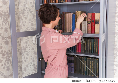 Young caucasian young woman with brunette hair taking boor for reading on free time standing near bookcase, positive girl selecting literature taking novels from shelves in library. Stay home. 64878839