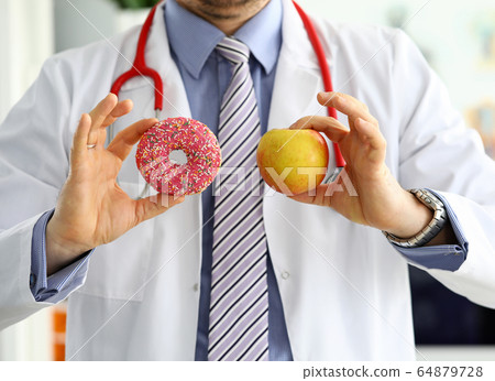 Doctor in office holding in hand pink doughnut vs red apple Doctor in office holding in hand pink doughnut vs red apple 64879728