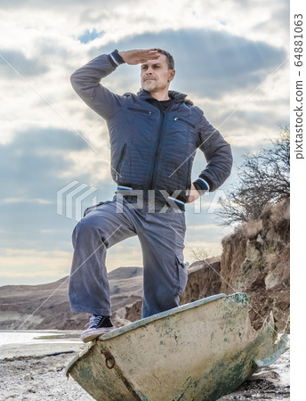 man in black stands in a broken boat on the beach and carefully looks into the distance man in black stands in a broken boat on the beach and carefully looks into the distance 64881063
