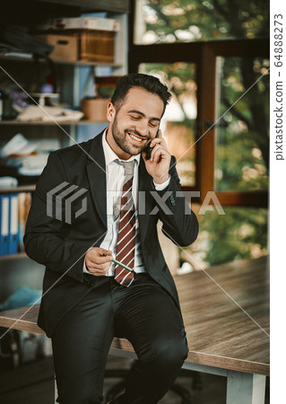 Cheerful Happy Businessman Talks With Mobile Phone Sitting On Desk Cheerful Happy Businessman Talks With Mobile Phone Sitting On Desk 64888273
